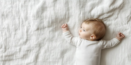 Baby girl is lying on a white blanket, wearing a white bodysuit, with her arms outstretchedの素材