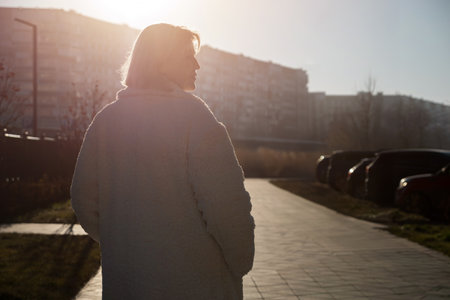 Blonde woman wearing a white coat walking away on a city street at sunset, with a blurred person pulling a suitcase in the backgroundの写真素材