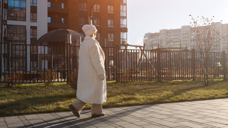 Senior woman enjoying a leisurely stroll on a paved walkway near modern apartment buildings, embracing the warmth of a sunny dayの写真素材