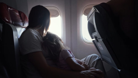 Happy woman plays with little daughter in cabin of airplane. Child admiringly follows scenery...