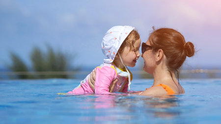 Loving mother and daughter sharing joyful moments while playing together in sparkling blue swimming pool under sunny summer skyの写真素材