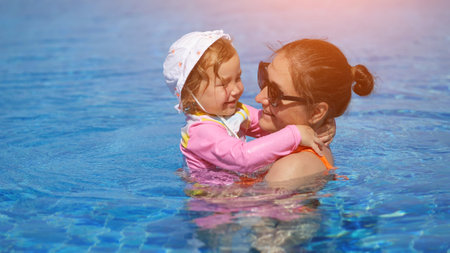 Mother cradles daughter in swimming pool and smiles reflecting love along with fun. Little girl hat and sleeves keep child safe from sunburnの写真素材