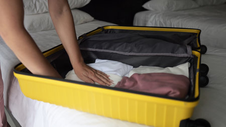 Young traveler neatly organizing yellow suitcase with clothing while preparing for upcoming vacation inside cozy hotel bedroomの写真素材