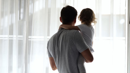Father holds his daughter in his arms as they gaze out of a window, enjoying the view togetherの写真素材