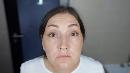 Close-up of a woman studying her forehead wrinkles in a bathroom mirror, expressing concern over aging skin and beauty routinesの写真素材