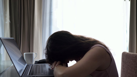 Woman rests head on hands visibly overwhelmed. Lady freelancer takes sip of tea seeking brief moment of relief. Small break seems to ease tensionの写真素材