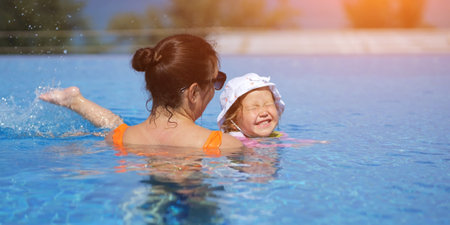 Mother gently supports daughter in swimming pool teaching to swim. Laughing toddler girl practices strokes gaining confidence with every moveの写真素材