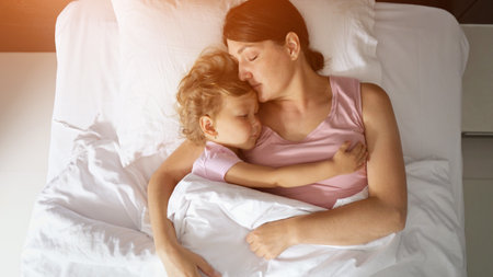 Loving mother and young daughter peacefully sleeping, closely embracing while resting on soft white hotel bedsheets during peaceful family vacation momentの写真素材