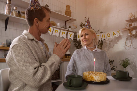 Happy mature couple wearing party hats, celebrating birthday with cake and tea, enjoying a cozy moment together in their kitchenの写真素材