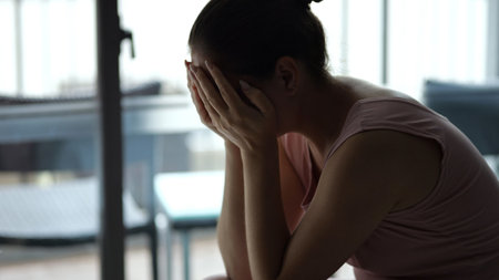 Distraught woman sitting alone on hotel bed, covering face and expressing deep emotional pain during challenging travel experienceの写真素材