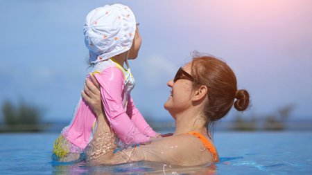 Happy mother lifting her daughter while swimming and playing in a pool during a sunny dayの写真素材