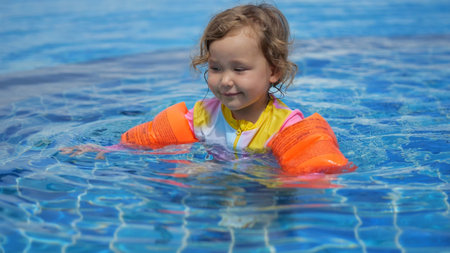 Cute toddler enjoying playful moments while swimming and splashing in a pool, wearing bright...