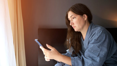 Female tourist sits on bed edge focused on phone in hotel apartment. Luxurious surrounding...