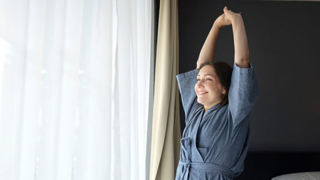 Woman stretches stepping to window soaking morning tranquility. Morning light bathes lady in soft glow and apartment embraces with peace and comfortの写真素材