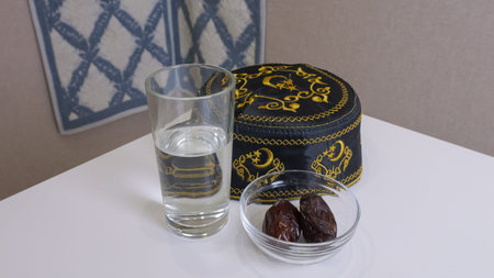 Glass Of Water With Dates, Embroidered Cap And Small Bowl On White Table, Clear Glass Reflecting...