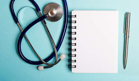 A stethoscope, a blank notepad and a pen lie on a light blue background. Top view close upの写真素材