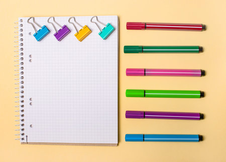 On a beige background there is a blank notebook, colored paper clips and various colored markers....