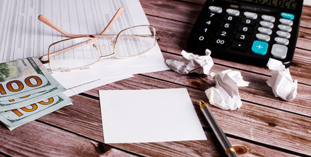On a wooden table, a calculator, cash, a pen with paper and gold-framed glasses. Business concept