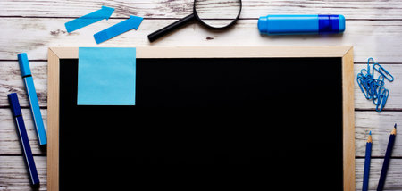 On a light wooden background, a black lettering board, blue and light blue markers, pens and pencils, a magnifying glass and paper clips with stickers. Place to insert text.の写真素材