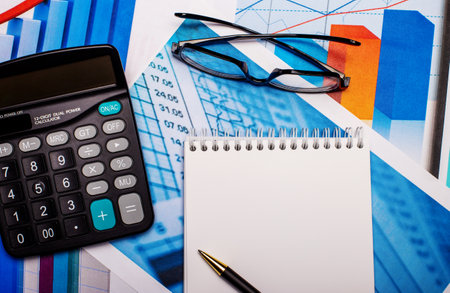 Glasses, a calculator, a blank notebook and a pen are on the bright diagrams. Workplace close upの写真素材