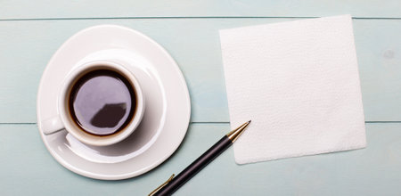 On a light blue wooden background, a white cup of coffee, an empty napkin for notes and a pen. Top view with copy spaceの写真素材