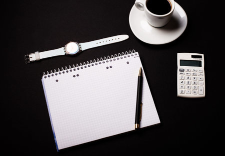 Coffee in a white cup, a wrist watch, a calculator and a blank open notebook on a black background....