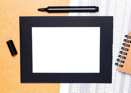 On a wooden table are a black open marker, a brown notepad and lst of paper in a black frame. Top...
