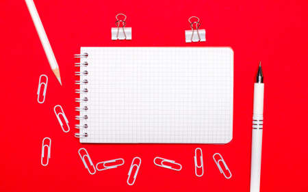 On a red background, a white pen, white paper clips, a white pencil and a blank notebook. Top view with copy spaceの写真素材