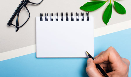 Glasses, a plant, a blank white notepad and a man's hand with a ballpoint pen lie on a gray and...