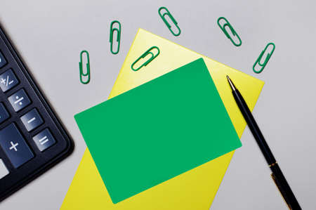 There is a calculator on a gray background, green paper clips, and a green card on a yellow sheet for inserting text or illustrations. Template. Flat layの写真素材