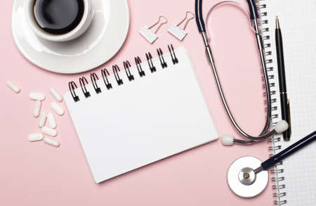 On a light pink background, a white cup of coffee, white pills, a stethoscope, a pen and a white notepad with a place to insert text or illustrations. Template. Medical conceptの写真素材