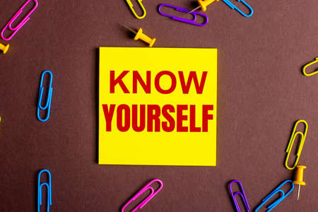 The words KNOW YOURSELF is written in red on a yellow sticker on a brown background next to multi-colored paper clips.の写真素材