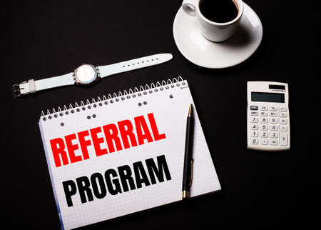 Coffee in a white cup, a wrist watch and a calculator on a black table. Nearby is a pen and a notebook with the text REFERRAL PROGRAMの写真素材