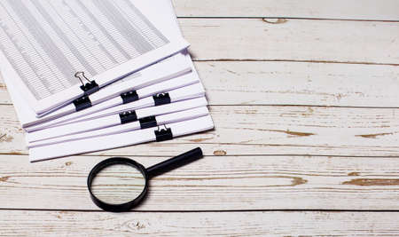 A stack of documents and a magnifying glass lie on a light wooden background. Place to insert text. Business conceptの写真素材