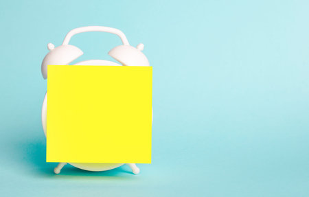 Against a light blue background, there is a white alarm clock with a yellow note sticker on it....