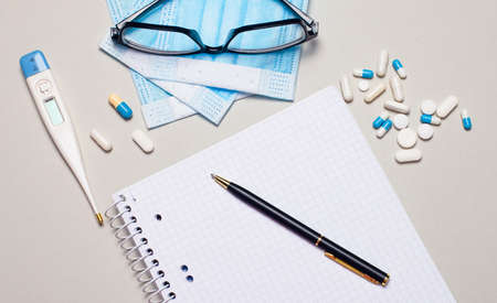 A face mask, pills, an electronic thermometer, glasses in a black rim, a notebook and a pen lie on a gray background. Medical conceptの写真素材