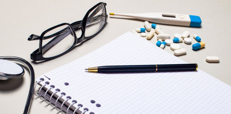 On a gray background lie a stethoscope, pills, an electronic thermometer, black-framed glasses, a notebook and a pen. Medical conceptの写真素材