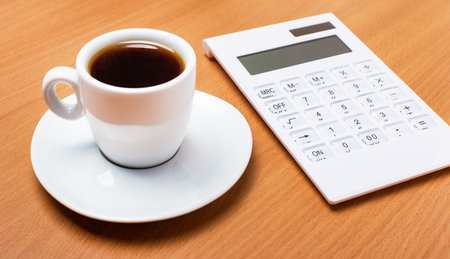 There is a white cup with coffee and a white calculator on a wooden table. Business concept