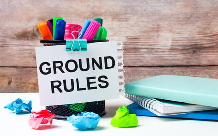 On the desktop and against the background of a wooden wall, there is a stand with multi-colored felt-tip pens, bright pieces of paper and a sheet of paper with the text GROUND RULESの写真素材