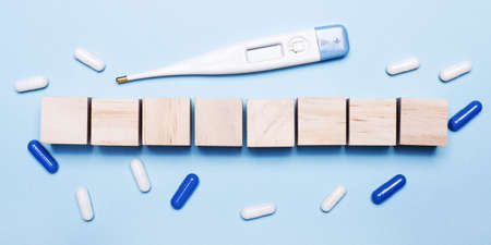 On a light blue background, an electronic thermometer, white and blue pills, empty wooden cubes with a place to insert text. Template. Medical conceptの写真素材