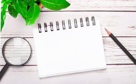 On a light wooden background, a magnifying glass, a pencil, a green plant and a white notebook with...