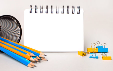 On a light background, yellow and light blue pencils and paper clips and a white blank notebook with...
