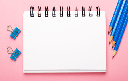 On a light pink background, light blue pencils and paper clips and a white blank notebook with space...