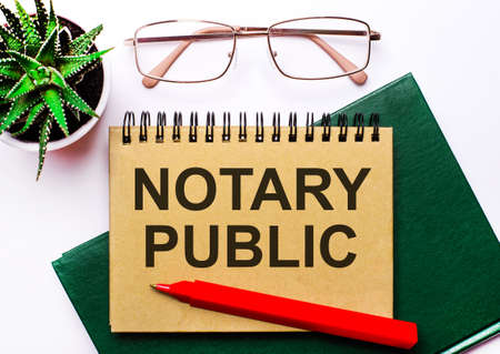 On a light background, gold-framed glasses, a flower in a pot, a green notebook, a red pen and a brown notebook with the text NOTARY PUBLIC. Business conceptの写真素材