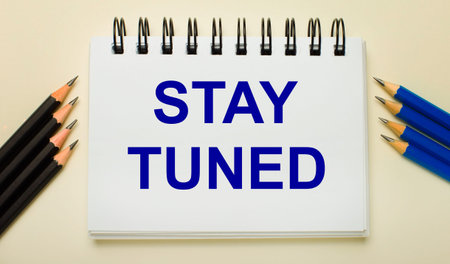 On a light background, a white notebook with the text STAY TUNED and black and blue pencils on the...