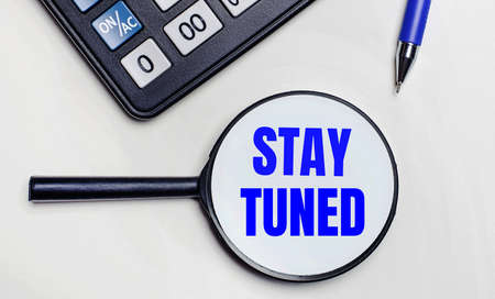 On a light background, a black calculator, a blue pen and a magnifying glass with text inside the word STAY TUNED. View from aboveの写真素材