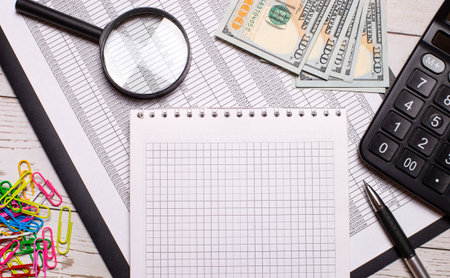 On the office desk is a notebook with a pen, calculator, dollars, multi-colored paper clips and a...