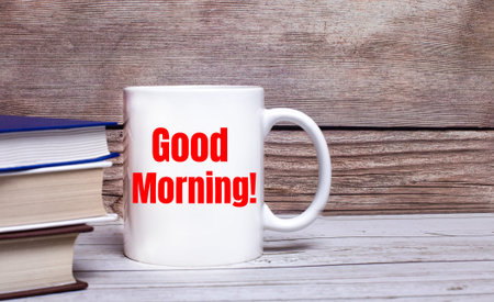 On a wooden background, there are books and a white cup with the text GOOD MORNING on it