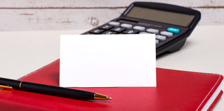 On the office table there is a burgundy notebook, a calculator, a pen and a white blank card with a...