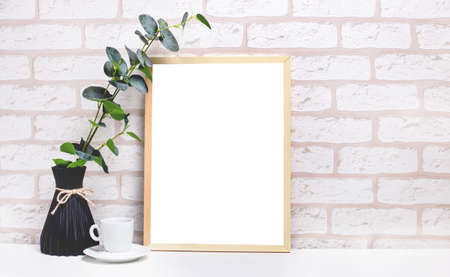 On a white table against the background of a light brick wall, there is a branch of eucalyptus in a dark vase, a white cup and a light wooden frame with a place to insert tex. Home office interior. Copy spaceの写真素材